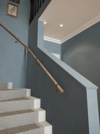 Angled view of Oversley Walnut Single Winder Handrail with Loxley Antique Brass brackets beside carpeted stairs against a blue-grey wall.